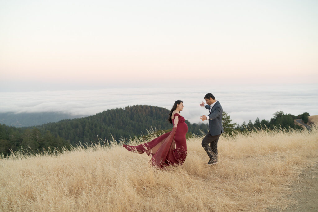 Husband and pregnant wife spread their arms in Mt. Tam for a hug.