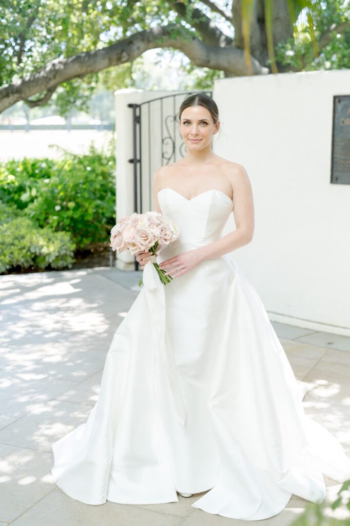 Bride wears a pristine white wedding gown with a long train and holds a fresly cut bouquet of roses.