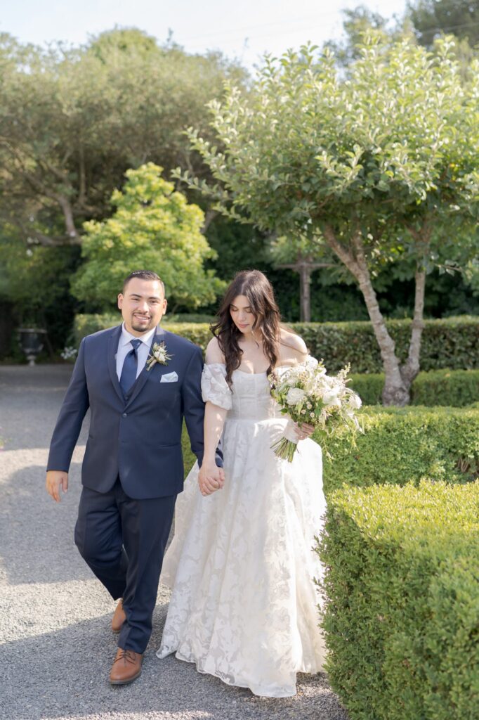 Newly married bride and groom hold hands and walk through neatly trimmed shrubs.