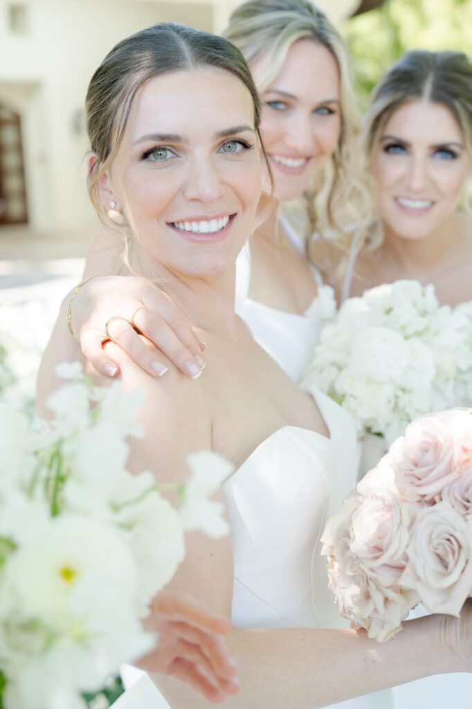 Bride and her bridesmaids hold rose bouquets and hug each other.