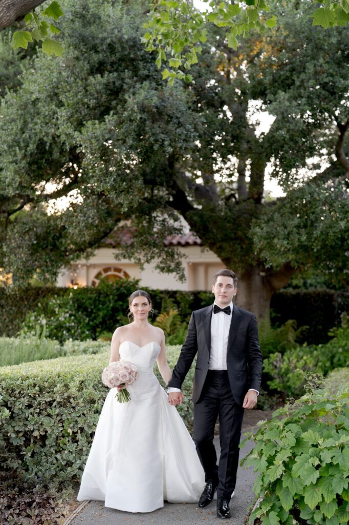 Groom in suit and bow tie and bride in wedding gown hold hands and walk through a beautiful garden.