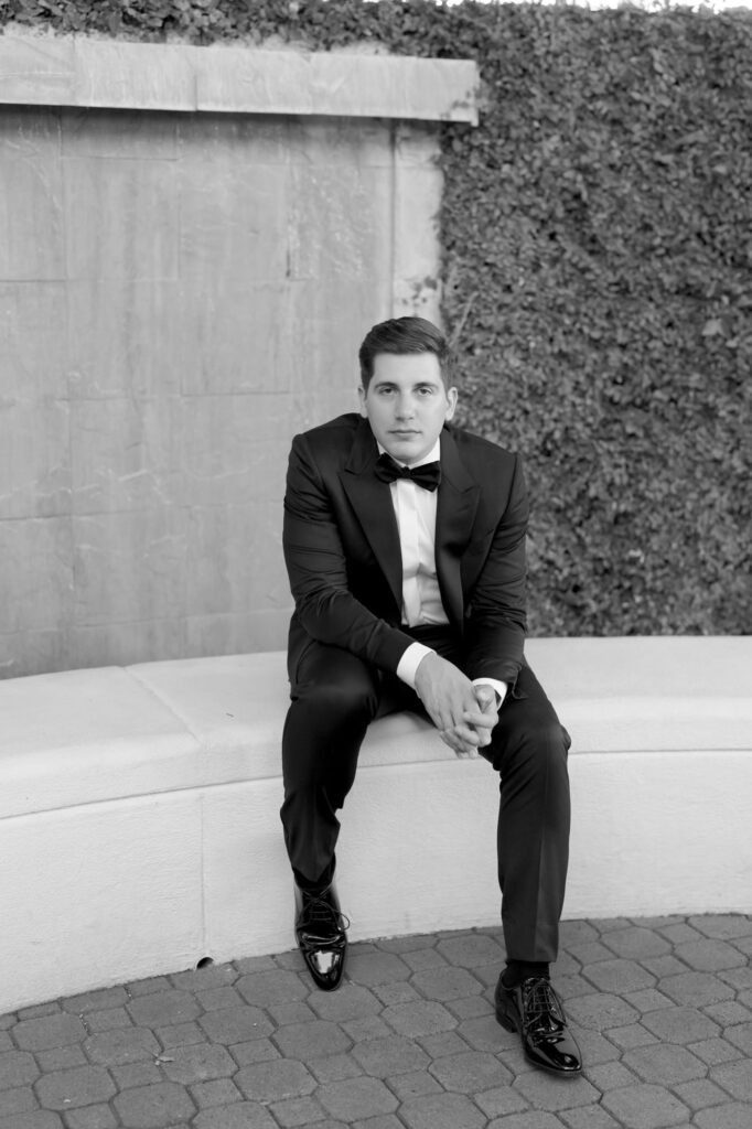 Handsome groom joins his hands and squats at a stone sitting arrangement.