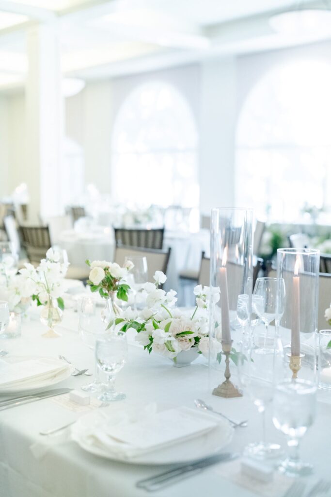 Long candles inside glass containers and flower vases placed alongside drinking glasses and cutlery on a table with white tablecloth.