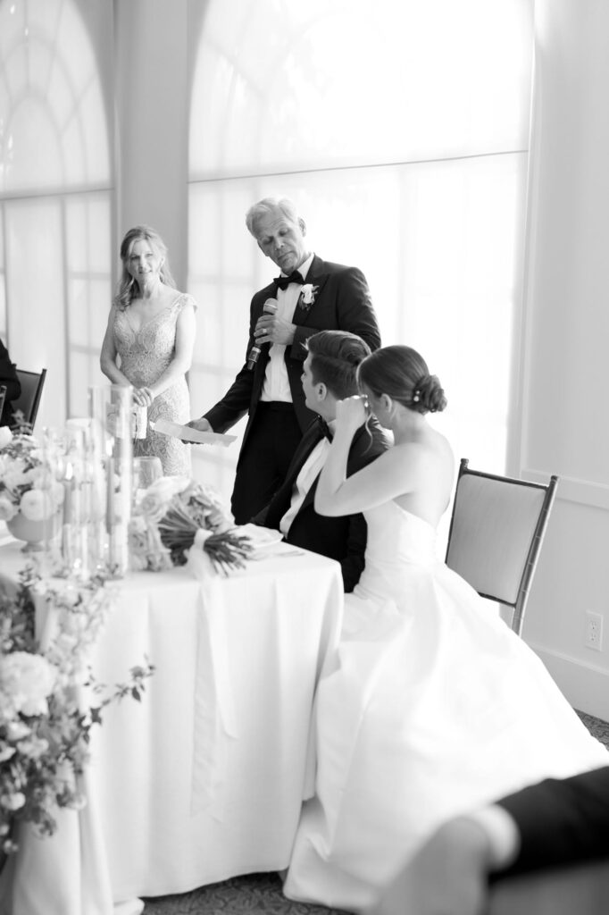 Wedding guest delivers his speech and congratulates the bride and groom who sit beside at a table and listen to him.