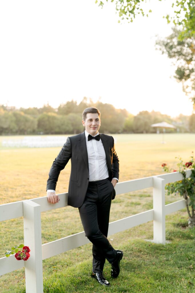 Impressively dressed groom leans on a white fence at Menlo Circus Club.