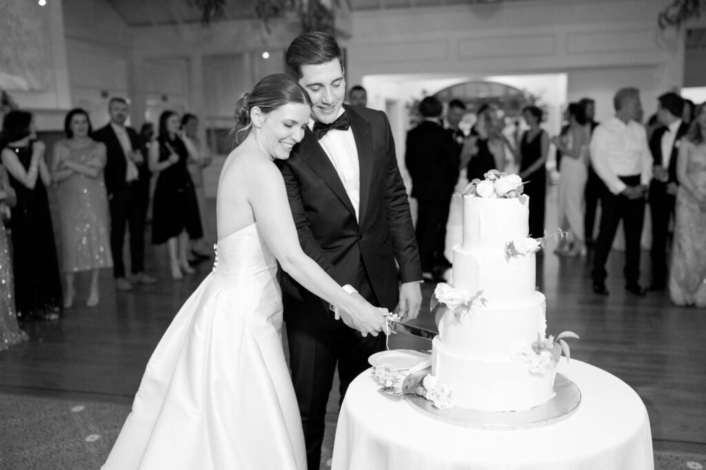 Bride and groom cut their wedding cake while guests watch from behind.