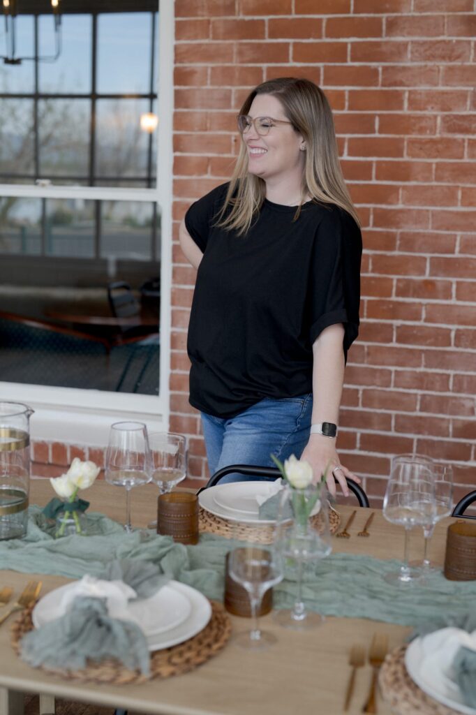 Lady in white t-shirt and blue jeans smiles while standing behind a dining table.